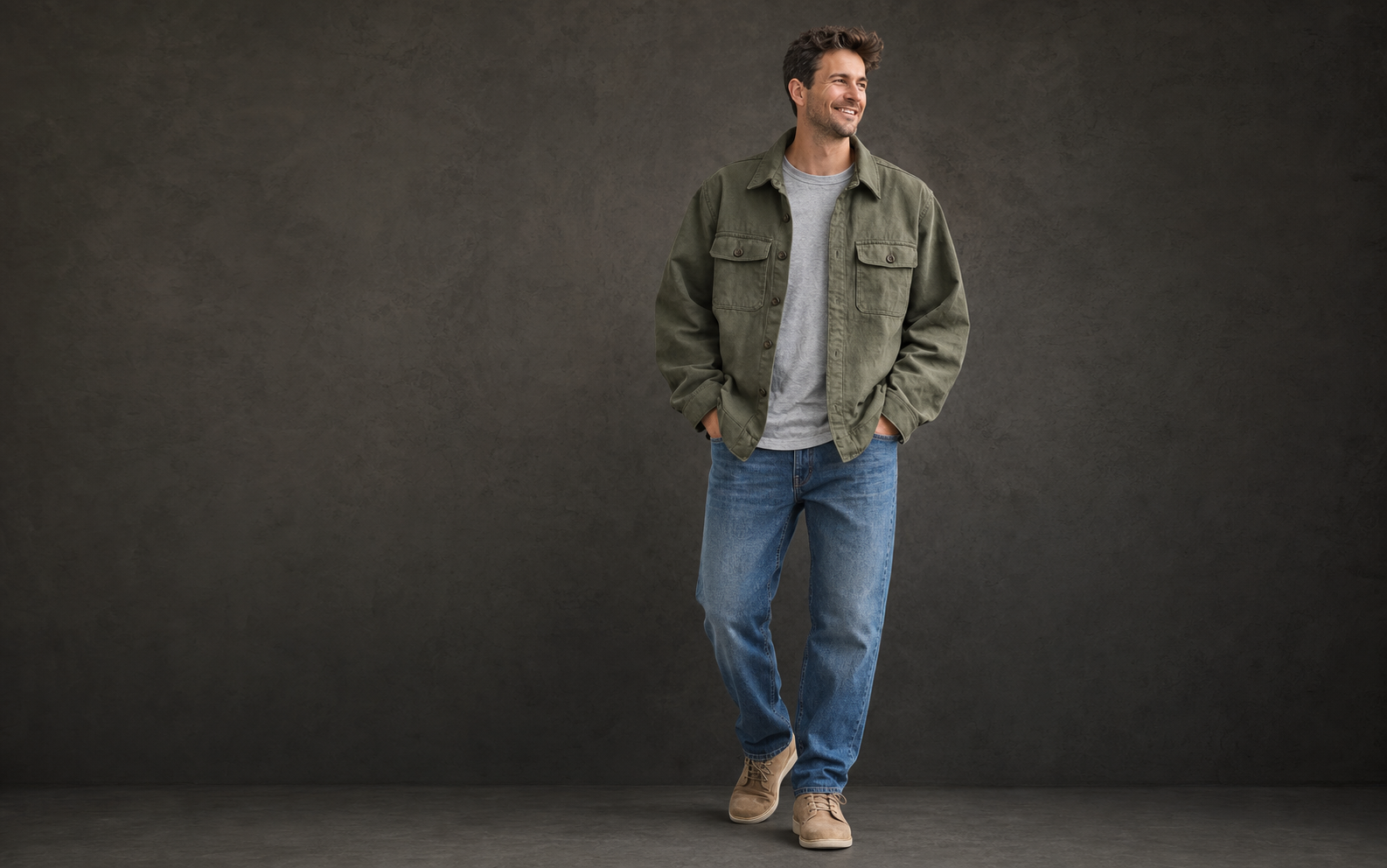 A man wearing an olive overshirt, gray T-shirt, blue jeans, and tan shoes in a dark gray studio